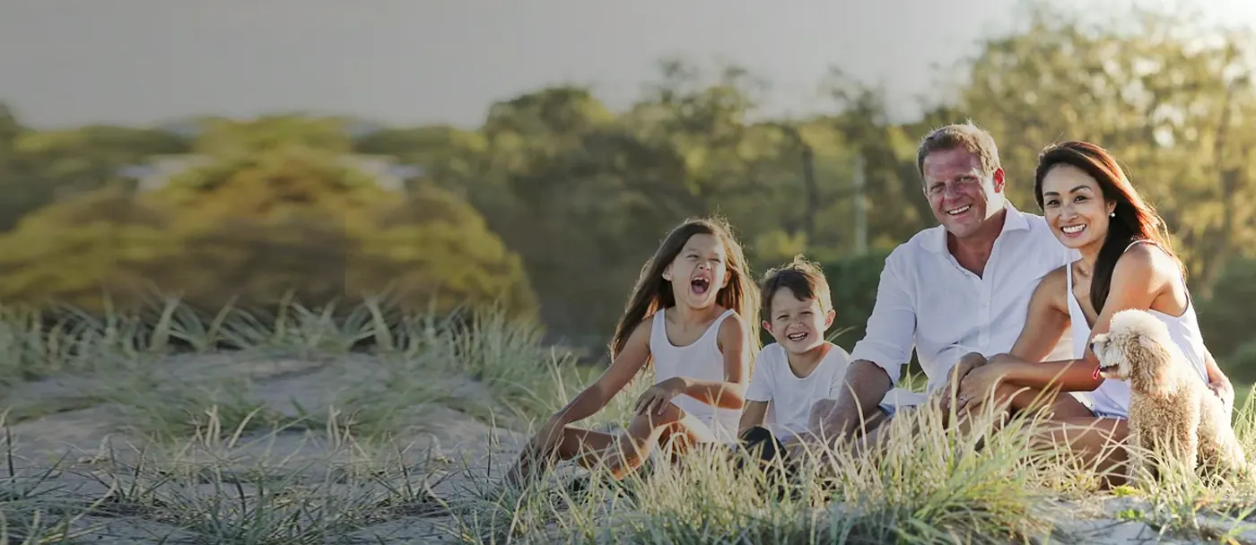Família sorrindo sentada na grama em um fim de tarde, com o cachorro ao lado